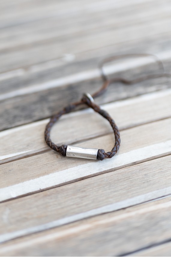 Pulsera Nómada de cuero marrón trenzado con pieza tubular de metal plateado y cierre ajustable.