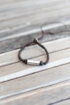 Pulsera Nómada de cuero marrón trenzado con pieza tubular de metal plateado y cierre ajustable.