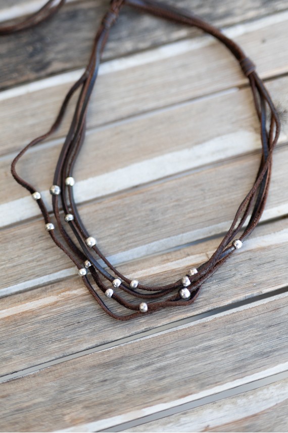 Detalle de collar de cuero marrón con múltiples tiras y pequeñas bolas de zamak distribuidas a distintas alturas.